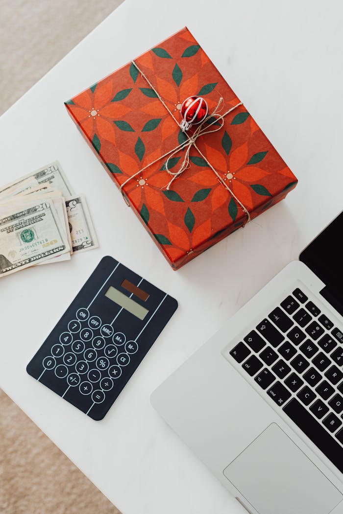 A beautifully wrapped gift box, cash, calculator, and laptop on a white table, symbolizing holiday expenses.